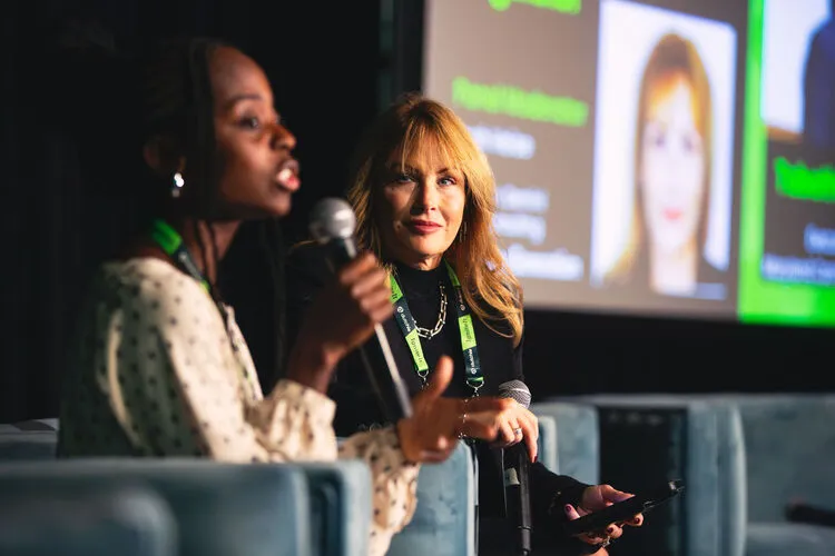 Two women on stage at a conference. One speaks into a microphone, while the other listens attentively, holding a phone. A presentation screen is visible behind them