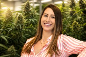 Portrait of Jessica Ferranti smiling in a commercial indoor cannabis cultivation facility, with mature flowering cannabis plants filling the background under grow lights.