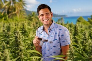 Khalifa Kush CEO DJ Saul smiling in a lush tropical cannabis field with flowering plants, palm trees, and a coastal landscape in the background.